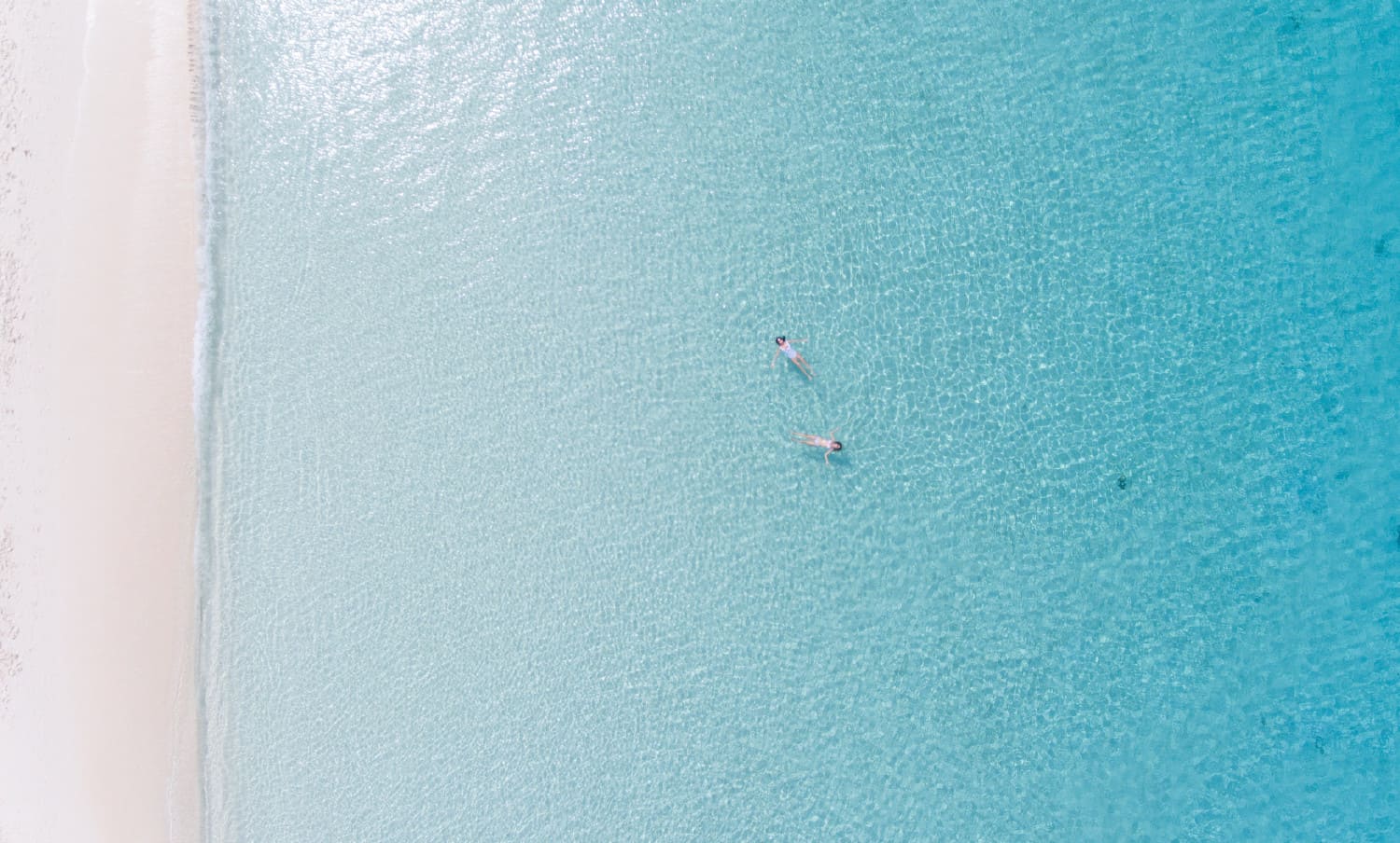 2 women floating in a crystal blue sea and white sand beach